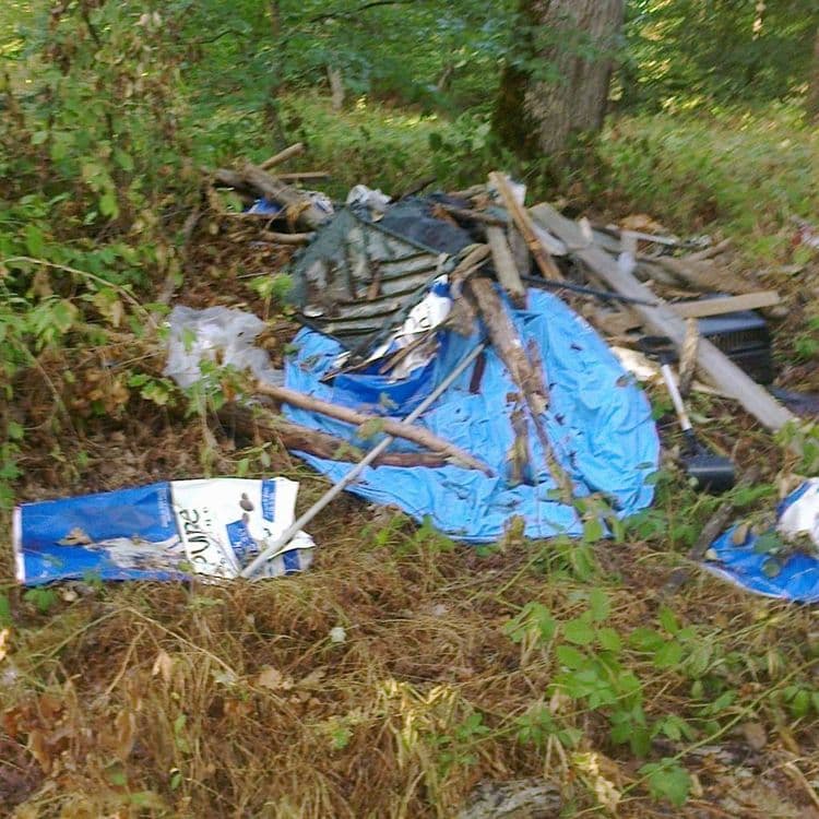 Dépôt de déchets à l'entrée d'un sentier. Présence de nombreux sacs vides de croquettes pour chien, d'une piscine en plastique et de divers objets. 
