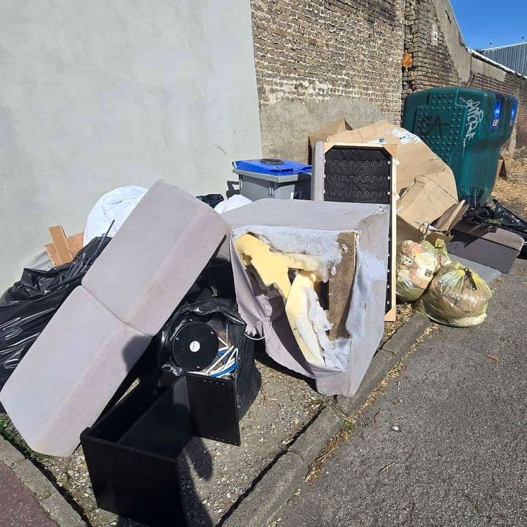Voici une estimation des déchets présents sur les lieux :

On observe un dépôt sauvage composé principalement de déchets suivants :

*   **Mobilier rembourré (canapé/fauteuil)** : Présence d'éléments de canapé démantelés, incluant l'armature, les coussins (dont certains sont déchirés laissant apparaître la mousse) et des ressorts. On peut estimer le poids total de ces éléments à environ 30 kg. La quantité est de deux assises et un dossier.

*   **Sacs poubelles** : Deux sacs poubelles semblent contenir des ordures ménagères. Le poids de chaque sac est estimé à environ 10 kg, soit 20 kg au total.

*   **Cartons** : Divers cartons de différentes tailles sont visibles, principalement aplatis. Le poids total de ces cartons est estimé à environ 5 kg. La quantité est estimée à 4 cartons.

*   **Déchets divers en sac noir** : Un sac noir opaque laisse présumer la présence de déchets divers non identifiables sans ouverture. Le poids estimé est de 10 kg.

*   **Haut-parleur** : un haut parleur noir d'environ 5kg est présent

*   **Meuble noir en bois** : Un meuble en bois noir est présent le poids est estimé à 10kg

*   **Poubelle bleue** : Une poubelle bleue de tri sélectif d'environ 1kg est présente.