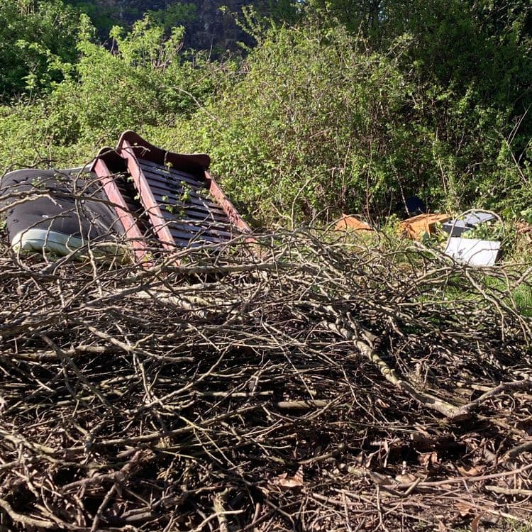 Voici l'analyse des déchets présents sur l'image :

*   **Mobilier** : Un sommier de lit avec matelas est visible. Le poids estimé pour cet ensemble est d'environ 20 kg. Il semble qu'il n'y ait qu'un seul ensemble de ce type. Une chaise de jardin en plastique est également présente, pesant environ 2 kg.

*   **Déchets Divers** : Des déchets divers sont visibles parmi les branchages, tels que des restes de meubles en bois et en métal, ainsi qu'un récipient en plastique. Le poids estimé est de 10 kg.

*   **Déchets verts** : Une importante quantité de branchages coupés est visible au premier plan. Le poids estimé de ces déchets verts est d'environ 30 kg.
