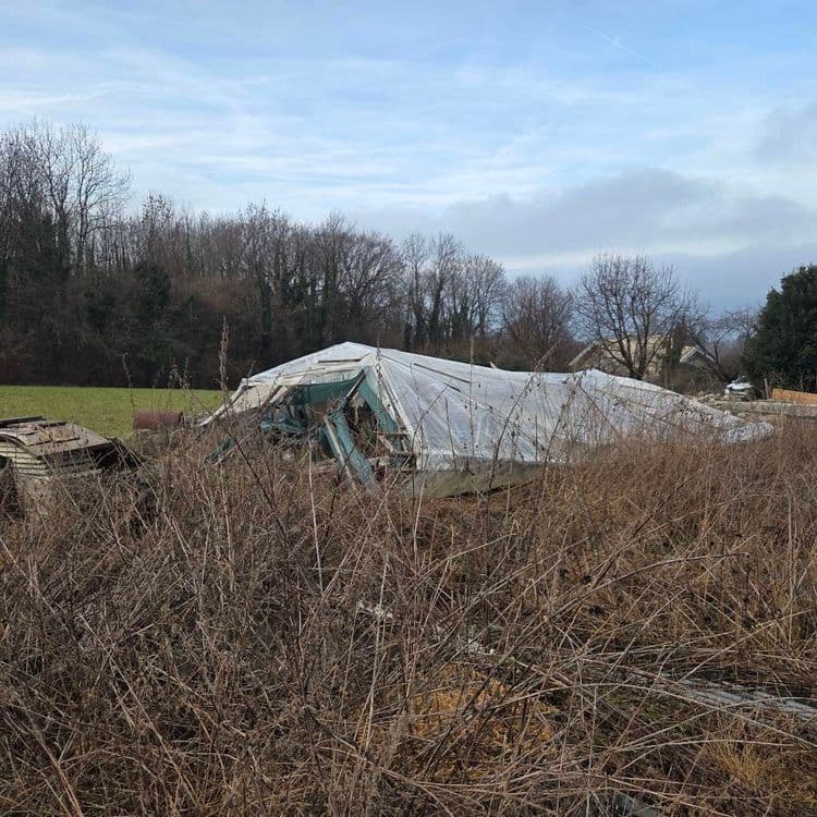 Sur la base des éléments observés, voici une estimation des déchets présents sur le site :

*   **Débris de serre :** Structure en bois et armature métallique d'une serre détruite, recouverte d'une bâche plastique. Estimation : 200 kg, 1 unité.
*   **Bois de construction :** Planches, panneaux de bois et éléments de coffrage. Estimation : 150 kg, environ 1 mètre cube.
*   **Divers éléments métalliques :** Fragments de métal, ferraille et pièces mécaniques non identifiées. Estimation : 80 kg, quantité variable.
*   **Bâche plastique :** Restes de la couverture de la serre, probablement en polyéthylène. Estimation : 10 kg, 1 unité.
*   **Remorque :** Une remorque est présente. Estimation : 150 kg, 1 unité.
*   **Fût bleu :** Un fût bleu en plastique (probablement 200 litres). Estimation : 10kg, 1 unité
*   **Citerne métallique :** Une citerne métallique. Estimation : 80 kg, 1 unité.

Ces estimations sont données à titre indicatif et peuvent varier en fonction de la nature exacte des matériaux et de leur état de dégradation. Une inspection sur place serait nécessaire pour une évaluation plus précise.
