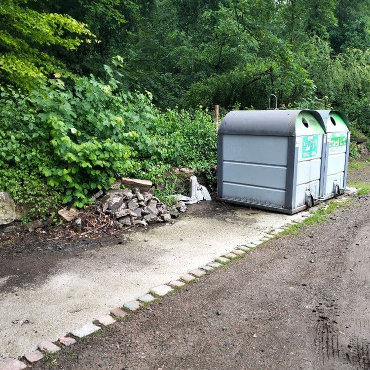Sur parking public en bordure de forêt, gravats de chantier et un aspirateur à côté d'un container à verre 