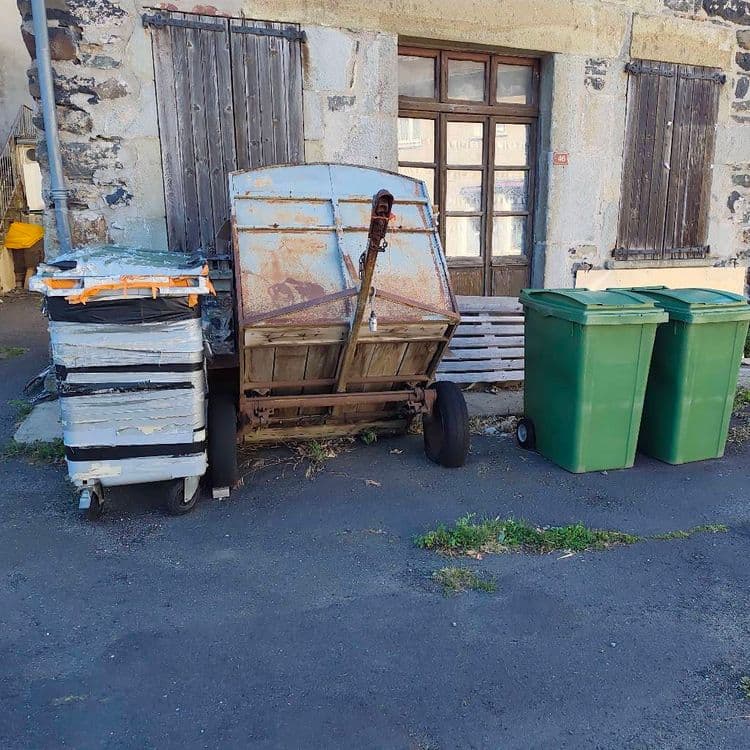 Voici les éléments identifiés sur le site :

*   **Une remorque ancienne** : Composée de ferraille et de bois, fortement corrodée.
    *   Quantité : 1 unité.
    *   Poids estimé : 200 kilogrammes.

*   **Un empilement de bacs ou tiroirs divers** : Ces objets, vraisemblablement en plastique ou métal léger, sont empilés sur un petit chariot et maintenus par des sangles et du ruban adhésif.
    *   Quantité : 1 ensemble (environ 6 à 8 éléments empilés sur un chariot).
    *   Poids estimé : 60 kilogrammes.

*   **Des éléments en bois** : Il s'agit de planches ou de parties de palettes en bois, visibles derrière la remorque et les bacs de collecte.
    *   Quantité : L'équivalent d'environ 1 à 2 petites palettes.
    *   Poids estimé : 30 kilogrammes.

Deux bacs roulants de collecte de couleur verte sont également présents sur le site. Ils semblent être des équipements de collecte standard.