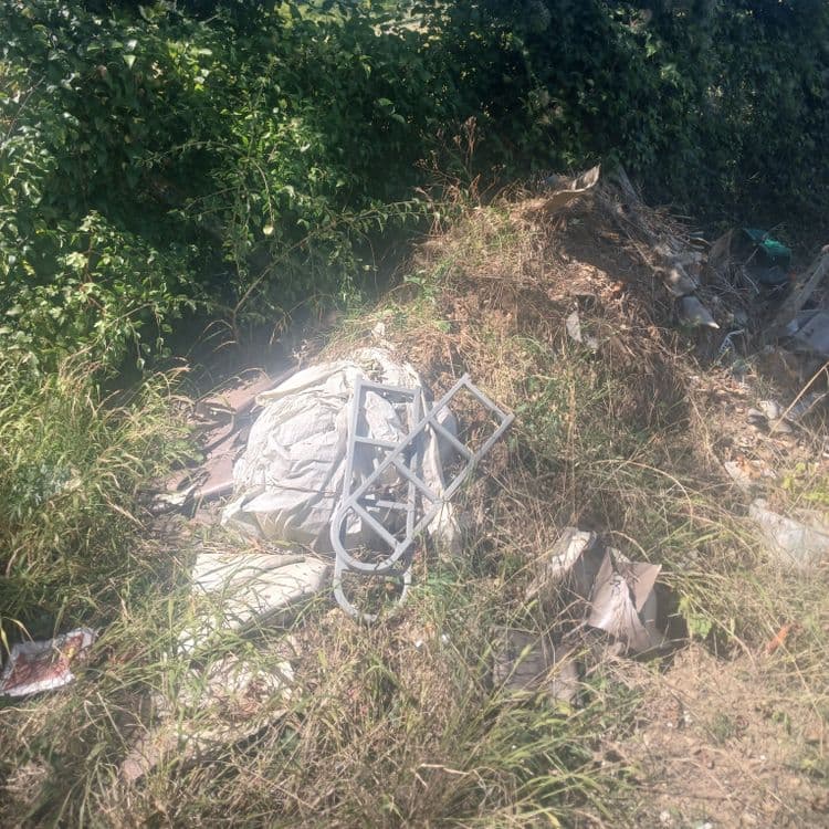 Voici l'analyse des déchets présents sur l'image :

*   **Déchets d'emballage (plastique et papier)** : On observe des sacs en papier et des fragments de plastique, probablement issus d'emballages divers. Le poids total estimé pour ce type de déchets est d'environ 2 kg, avec une quantité estimée à 5-7 éléments distincts.

*   **Déchets de construction/rénovation** : Des fragments de matériaux de construction, potentiellement des restes de plâtre ou dalles en béton, sont visibles. Le poids de ces déchets est estimé à 15 kg, avec une quantité estimée à 3-4 morceaux.

*   **Objets en plastique divers** : Une structure en plastique gris, dont la fonction initiale est difficile à déterminer, est présente. Son poids est estimé à 1 kg, avec une quantité de 1 objet.

*   **Déchets végétaux secs** : Une accumulation de branches sèches et d'herbes coupées est visible. Le poids total de ces déchets est estimé à 5 kg, avec une quantité importante en volume.

*   **Autres déchets non identifiables** : Quelques petits déchets épars, dont la nature précise est difficile à déterminer à partir de l'image, sont présents. Leur poids total est estimé à moins de 1 kg, avec une quantité de 2-3 éléments.