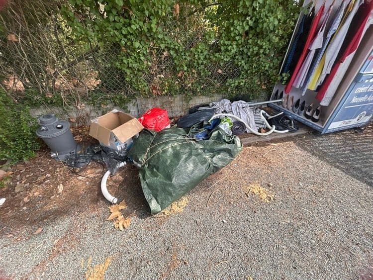 Les déchets abandonnés sont les suivants :

*   Un filtre ou une pompe de piscine, 1 unité, estimé à 20 kilogrammes.
*   Une boîte en carton vide, 1 unité, estimée à 0.5 kilogramme.
*   Des sacs plastiques remplis, dont 1 sac rouge et environ 3 sacs noirs, pour un total de 4 unités, estimés à 12.5 kilogrammes.
*   Une bâche verte nouée, probablement remplie de divers objets, 1 unité, estimée à 12 kilogrammes.
*   Des textiles et vêtements, comprenant une pile au sol et des articles sur le présentoir, pour un total d'environ 25 à 30 articles, estimés à 10 kilogrammes.
*   Une structure métallique de présentoir, 1 unité, estimée à 20 kilogrammes.
*   Des tuyaux flexibles, environ 3 sections, estimés à 4 kilogrammes.
*   Des chaussures, environ 4 paires, estimées à 2.5 kilogrammes.
*   Des déchets divers incluant des câbles et petits éléments plastiques, estimés à 2.5 kilogrammes.