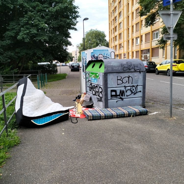 Matelas et déchets divers à côté de containers à verre et papier 